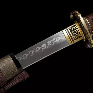 May include: Close-up of a Japanese katana sword. The blade is silver with a decorative pattern. The hilt is wrapped in brown fabric, and the guard is gold with a geometric design. The sword is set against a black background.