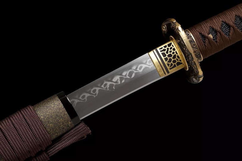 May include: Close-up of a Japanese katana sword. The blade is silver with a decorative pattern. The hilt is wrapped in brown fabric, and the guard is gold with a geometric design. The sword is set against a black background.