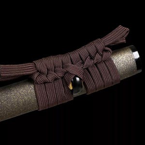 May include: Close-up of a katana handle with a dark brown cord wrap. The handle has a textured, metallic finish. The cord is intricately woven around the handle, with a knot detail near the top.