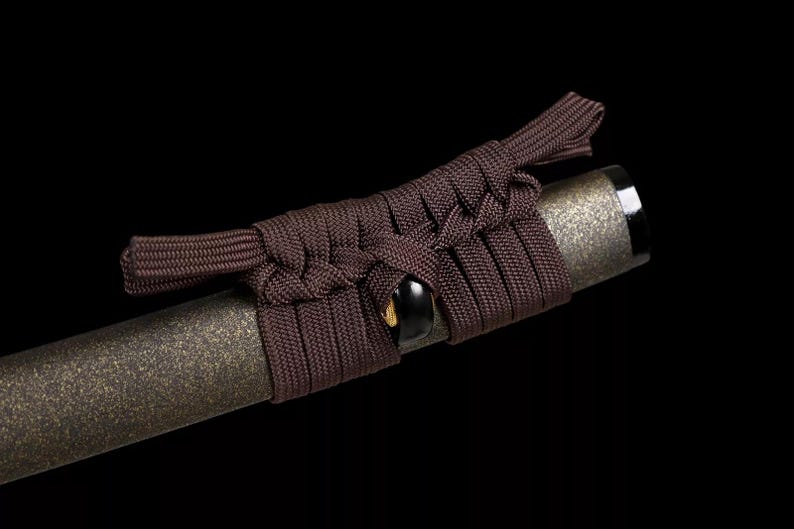 May include: Close-up of a katana handle with a dark brown cord wrap. The handle has a textured, metallic finish. The cord is intricately woven around the handle, with a knot detail near the top.