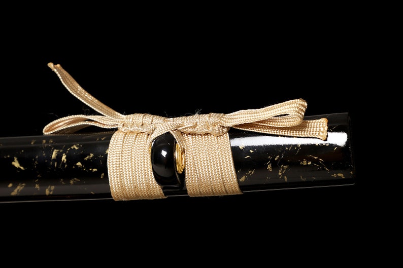 Pode incluir: Um close-up de uma bainha de katana preta e dourada com um cord&atilde;o bege enrolado ao redor.