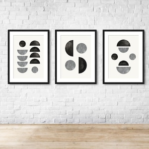 May include: Three framed abstract art prints. Each print features a black frame with a white background. The art includes geometric shapes in black, gray, and white, including circles and semi-circles. The prints are displayed on a white brick wall.