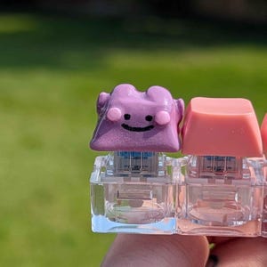 May include: A custom mechanical keyboard keycap shaped like a purple character with a hand-painted black smile and pink cheeks, mounted on a clear switch base next to a plain pink keycap, showcasing a glossy finish and detailed craftsmanship.