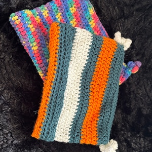 Op de afbeelding: Twee gehaakte tasjes met kleurrijke strepen. Eén tasje is regenboogkleurig en de andere is teal, wit en oranje.