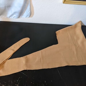 May include: A tan-colored medieval hood with a long, attached tie. The hood is made of a soft fabric and is laid flat on a dark surface. The hood's design includes a pointed top and a long, narrow tie.