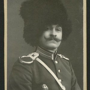 May include: A black and white portrait of a man in a military uniform. He is wearing a large, furry hat and has a mustache. The photo is from the early 1900s and has a vintage look.