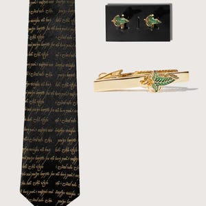 May include: A black tie with gold script and a matching tie clip, along with a set of cufflinks. The tie clip and cufflinks feature a gold leaf design with green accents. The tie has a repeating gold script pattern.