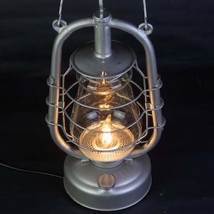 May include: A silver metal lantern with a glass shade and a light bulb inside. The lantern has a wire cage around the glass shade and a handle on top.