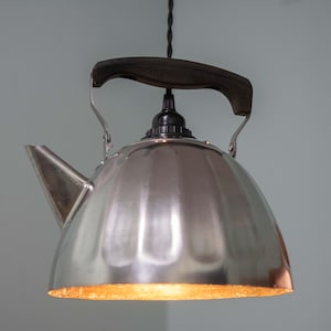 Może przedstawiać: Unikalna lampa wisząca wykonana z czajnika ze stali nierdzewnej. Lampa ma czarny uchwyt i czarny przewód elektryczny. Spód czajnika świeci ciepłym, zachęcającym światłem.