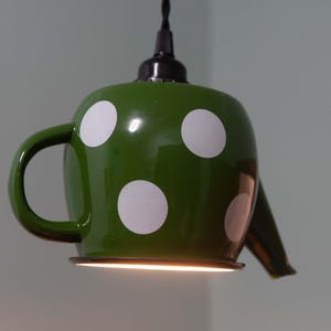 Può includere: Lampada a sospensione a forma di teiera verde con pois bianchi. Il lampadario ha un cavo nero e un manico laterale. Il beccuccio è sul lato destro e la base della lampada è illuminata.