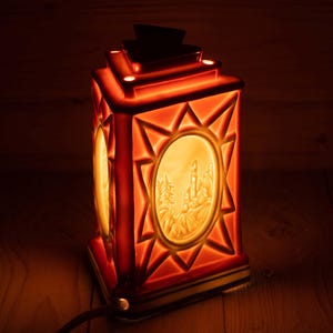 Könnte beinhalten: Eine rote Keramiklaterne mit sternförmigem Design und einer weißen Szene eines Dorfes im Inneren. Die Laterne ist beleuchtet und leuchtet.