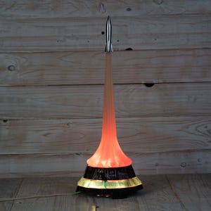 May include: A rocket-shaped lamp featuring a silver-toned metal rocket at the top, a translucent orange fluted column representing exhaust, and a black base decorated with white hieroglyphic-style symbols and a glowing yellow light ring at the bottom.