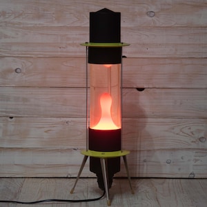 May include: A retro lava lamp with a black top and base, a clear glass cylinder, and a yellow accent ring. The lamp's interior features a glowing, orange lava shape. The lamp has a tripod base and a power cord.