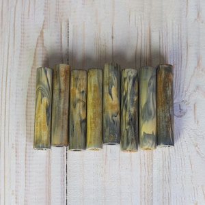 以下が含まれることがあります： 大理石模様の8つの円筒形のオブジェクトのコレクション。黄色、茶色、グレーの色合いがあり、ユニークな渦巻き模様を作り出しています。明るい色の木製の表面に配置されています。