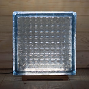 Peut inclure: Une lampe carrée en bloc de verre avec un cadre teinté de bleu et un intérieur texturé. La lampe repose sur une base en bois rectangulaire. Le cordon de la lampe est noir.