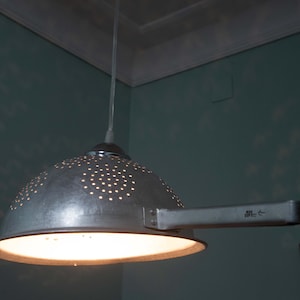 May include: A unique, industrial-style pendant light fixture. The lampshade is a repurposed metal colander with numerous small holes, suspended from the ceiling. The light fixture has a handle and emits a warm, inviting glow.