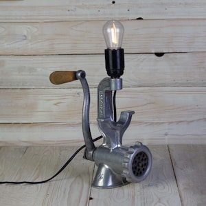 Può includere: Una lampada da tavolo in stile vintage realizzata da un macinaca carne riadattato. La lampada ha una finitura argentata e una maniglia in legno. È dotata di una presa nera con una lampadina trasparente.