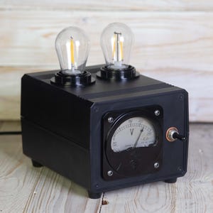 Puede incluir: Lámpara de mesa negra de estilo industrial con dos bombillas transparentes de estilo Edison. La lámpara cuenta con un voltímetro y un interruptor basculante. El diseño recuerda a equipos eléctricos antiguos.