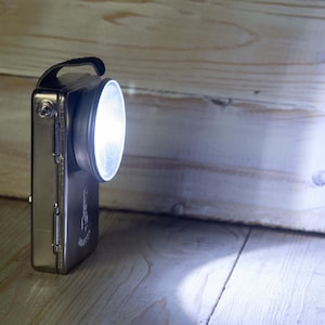 Puede incluir: Una linterna plateada de estilo vintage con un asa negra y una luz circular brillante. La linterna rectangular tiene un lado con bisagras y está colocada sobre una superficie de madera, proyectando un haz de luz.