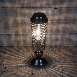 Puede incluir: Una lámpara de mesa decorativa con una pantalla cilíndrica texturizada y una base oscura. La lámpara emite un brillo cálido, proyectando sombras sobre la superficie de madera. La lámpara está enchufada y lista para usar.