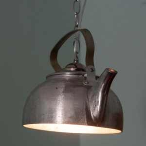 May include: A metal kettle-shaped pendant light, suspended by a chain. The light fitting has a handle and spout, with a warm glow emanating from the base. The fixture is designed to resemble a vintage kettle.