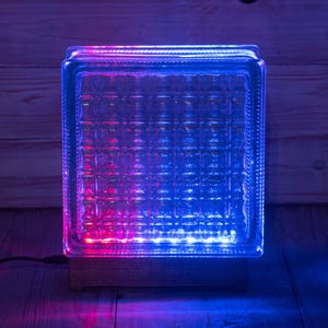 Può includere: Lampada a blocchi di vetro trasparente con motivo a griglia, illuminata da luci a LED rosa e blu. La lampada è posizionata su una base di legno ed è collegata a una fonte di alimentazione.