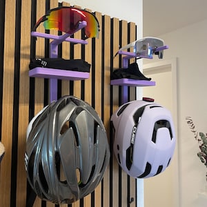 Könnte beinhalten: Ein Wand-Organizer für Sportausrüstung mit lila Regalen und Haken. Er hält eine Sonnenbrille, ein schwarzes Tuch und zwei Fahrradhelme, einen grauen und einen hellvioletten. Der Organizer ist an einer Holzlattenwand montiert.