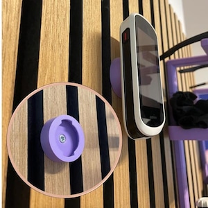 Può includere: Un dispositivo Garmin montato su una parete di legno a strisce nere. È visibile anche un supporto rotondo in legno con un supporto viola. Il dispositivo è bianco e nero con un pulsante rosso. Il muro ha un design moderno e minimalista.