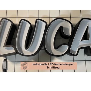 Könnte beinhalten: Ein schwarz-weißes LED-Namensschild, das den Namen "LUCA" buchstabiert. Das Schild besteht aus dicken, 3D-Buchstaben mit weißem Innenraum und schwarzem Außenraum. Das Schild ist mit weißen LED-Lichtern beleuchtet. Das Schild befindet sich auf einer weißen gekachelten Oberfläche.