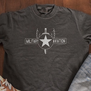 May include: Dark gray t-shirt with a white graphic that reads "MILITARY AVIATION". The graphic includes a star, wings, and a sword. The shirt is paired with blue jeans and sneakers.