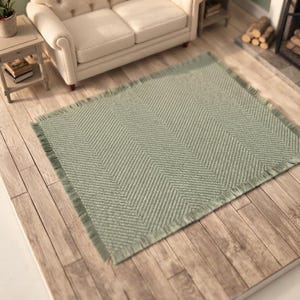 May include: A rectangular area rug in a soft sage green color featuring a prominent herringbone weave pattern. The rug is finished with a short, matching fringe along all four edges, creating a textured appearance across the entire surface.