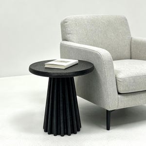 May include: A small, round black side table with a textured, ribbed base. A book rests on the table's surface. The table is next to a light gray armchair, creating a modern, minimalist aesthetic.
