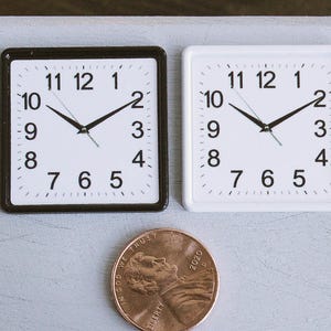 May include: Two small square clocks, one with a black frame and the other with a white frame, sit side-by-side. Both clocks have a white face with black numerals and hands. A penny is next to the clocks for size comparison.