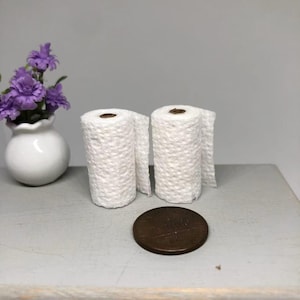 May include: Two miniature white toilet paper rolls with a penny next to them. The rolls are textured and partially unrolled. A small white vase with purple flowers is in the background.