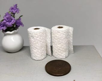 May include: Two miniature white toilet paper rolls with a penny next to them. The rolls are textured and partially unrolled. A small white vase with purple flowers is in the background.