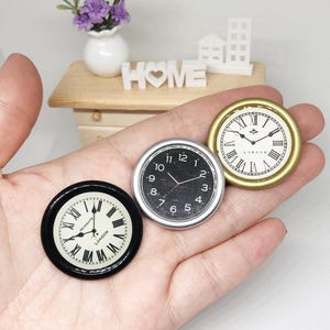 May include: Three miniature clock faces in a hand. One has a black frame with Roman numerals and the text "NEWGATE LONDON". Another has a silver frame with black numbers. The third has a gold frame with Roman numerals and the text "LONDON".