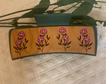 May include: A rectangular hair claw clip with a teal top and a wooden base. The base features four pink and yellow floral designs. The clip is designed to hold hair in place and is suitable for various hairstyles.