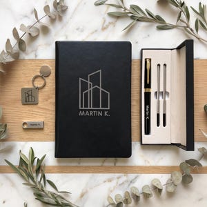 Pode incluir: Um caderno preto com um logótipo de edifício e o texto MARTIN K. gravado na capa, uma caneta preta a condizer com o nome Martin K. num estojo preto, um porta-chaves quadrado de metal com o mesmo logótipo e uma pequena pen USB retangular.