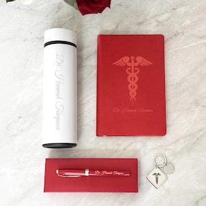 May include: A personalized gift set for a doctor, featuring a red notebook with a caduceus symbol and the name "Dr. Hannah Thompson", a white thermos with the same name, a red pen with the same name, and a silver keychain with a caduceus symbol.