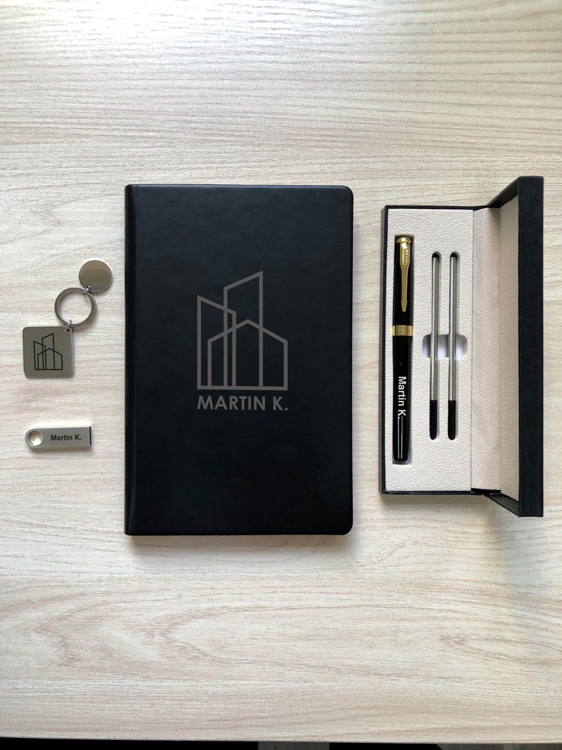 May include: A black notebook with a building logo and the text MARTIN K., a silver keychain with the same logo, a silver USB drive labeled MARTIN K., and a black pen with gold accents and the name MARTIN K. in a black presentation box.