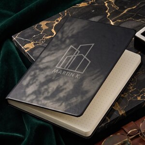 May include: A black notebook with a matte finish featuring a white line-art graphic of geometric buildings and the text MARTIN K. The notebook has cream-colored pages with a dot grid pattern and is partially open, revealing the interior paper texture.
