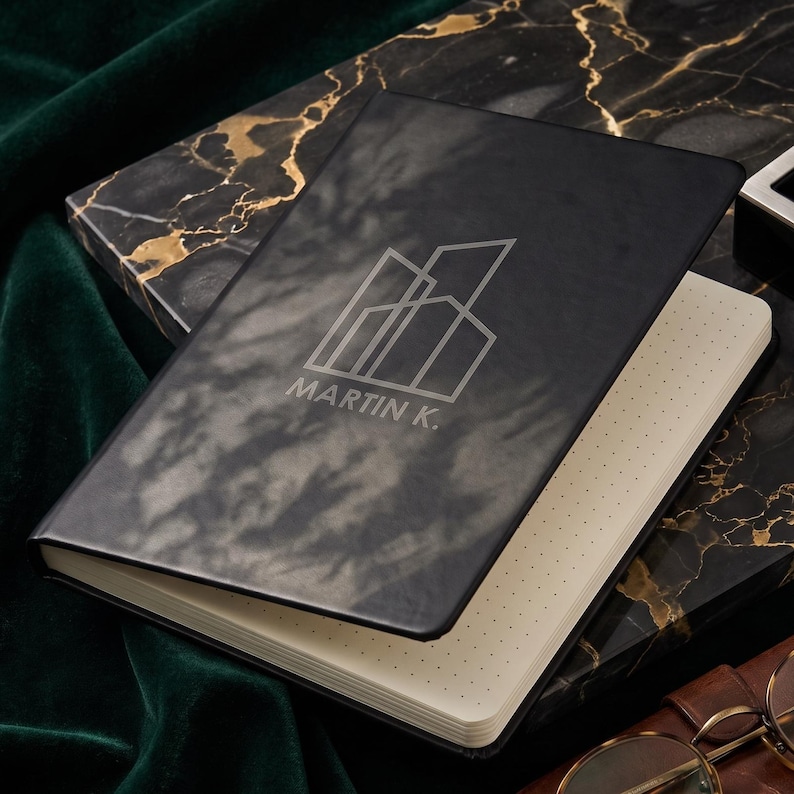 May include: A black notebook with a matte finish featuring a white line-art graphic of geometric buildings and the text MARTIN K. The notebook has cream-colored pages with a dot grid pattern and is partially open, revealing the interior paper texture.