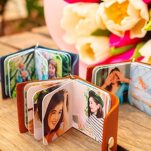 May include: Small photo albums with brown leather covers, each containing several photographs. The albums are open, revealing various images of people. A bouquet of pink and white tulips is in the background, on a wooden surface.