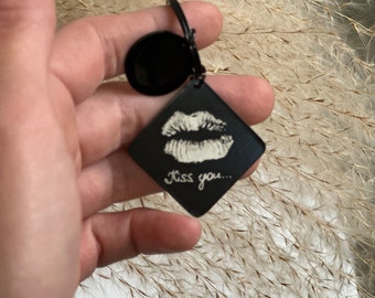 May include: Black keychain with a diamond-shaped charm featuring a white lip print and the text "Kiss you...". The keychain has a circular top and a black clasp.