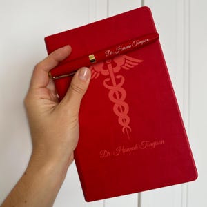 May include: Red leather journal with a gold pen and the text "Dr. Hannah Thompson" on the cover. The journal features a medical symbol, the caduceus, with a snake wrapped around a staff.