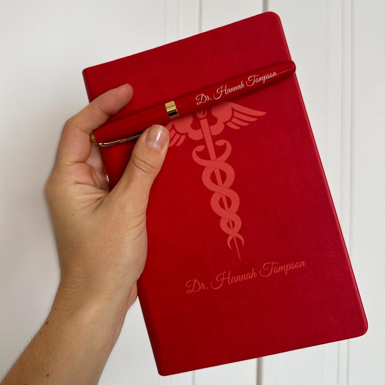 May include: Red leather journal with a gold pen and the text "Dr. Hannah Thompson" on the cover. The journal features a medical symbol, the caduceus, with a snake wrapped around a staff.