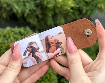 May include: A small, open photo album featuring two images of a person with a dachshund. The album is encased in a brown leather sleeve with a snap closure. The background is blurred green foliage.