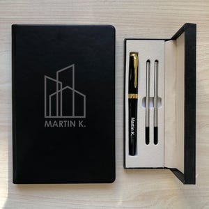 May include: A black notebook with a white line-art building logo and the text MARTIN K. next to a black gift box containing a black and gold pen with the text Martin K. and two silver-colored pen refills on a white interior tray.
