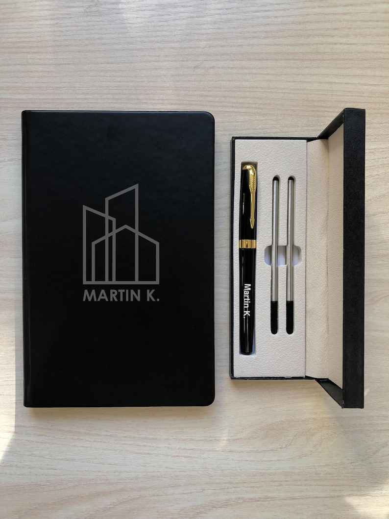 May include: A black notebook with a white line-art building logo and the text MARTIN K. next to a black gift box containing a black and gold pen with the text Martin K. and two silver-colored pen refills on a white interior tray.