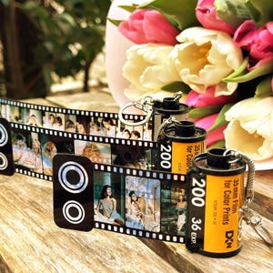 May include: A display of personalised film roll keyrings and a film strip with photos. The keyrings are black and yellow, with the text "200" and "36 EXP." printed on them. The film strip shows various photos. A bouquet of tulips is in the background.
